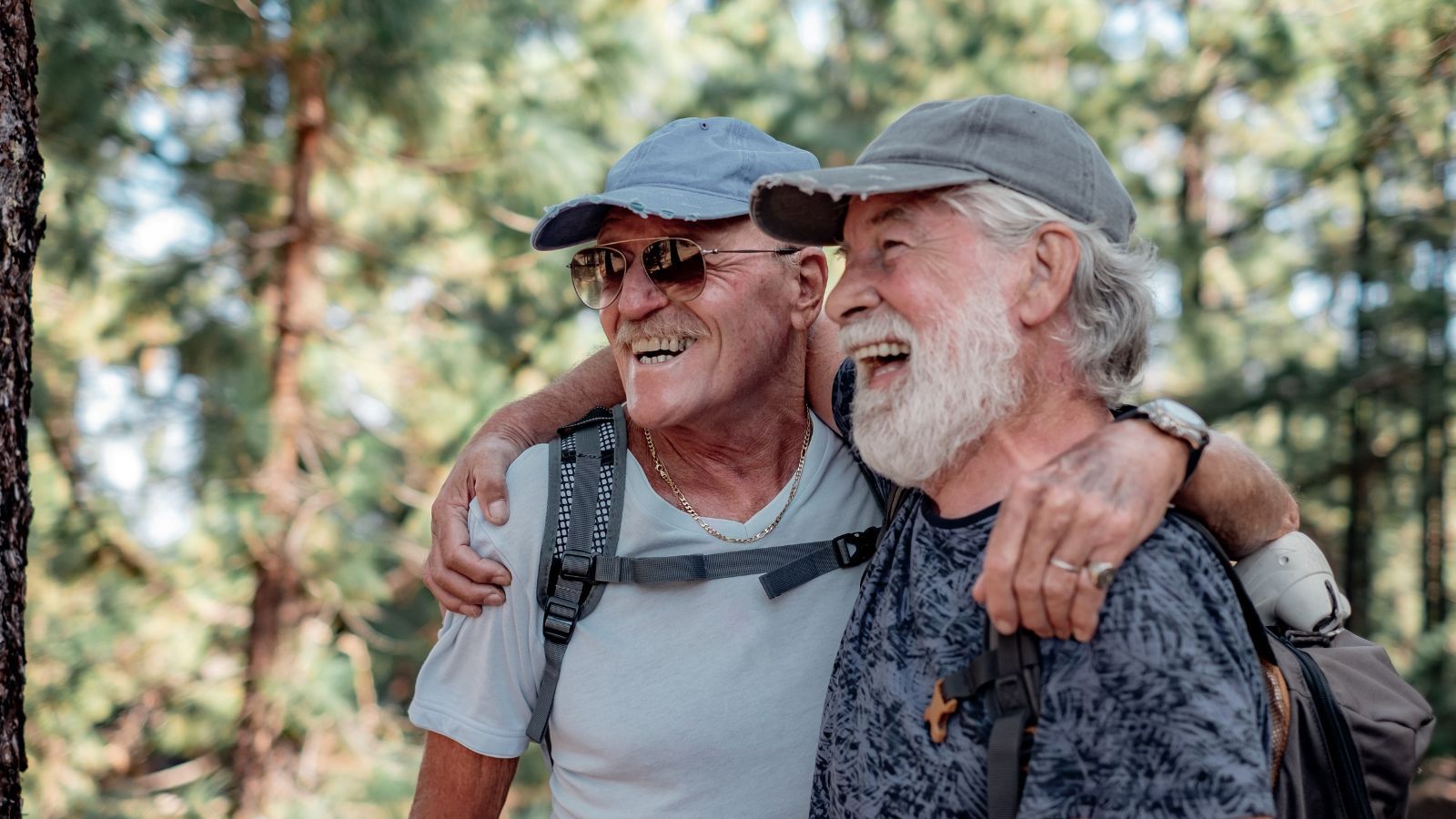 Happy couple of old active men with hat and backpack embrace and have fun in mountain hike in the woods appreciating adventure and freedom